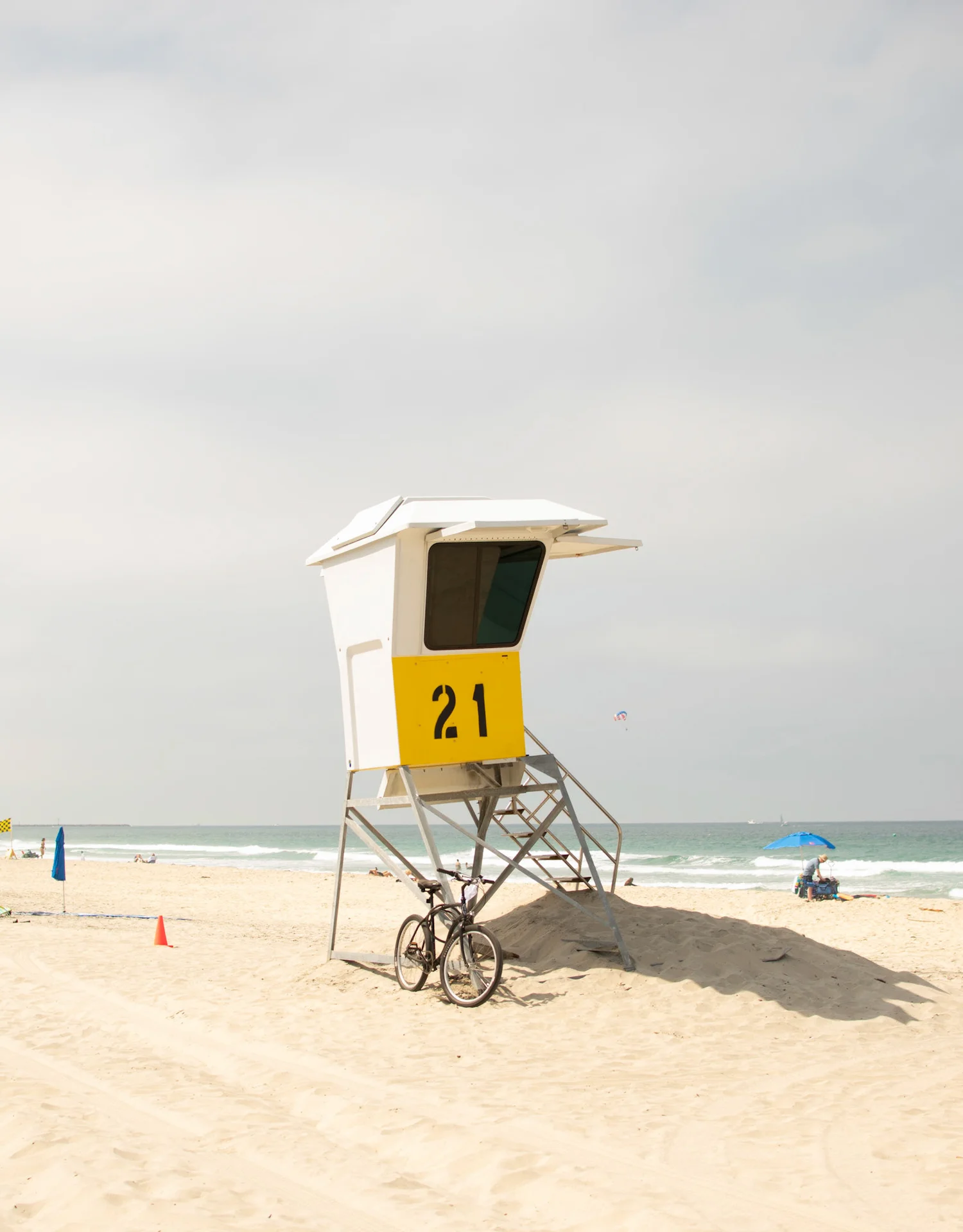 A solitary lifeguard tower stands on a sunlit beach, marked with the number 21, with a bicycle leaning nearby and a calm ocean stretching into the horizon.