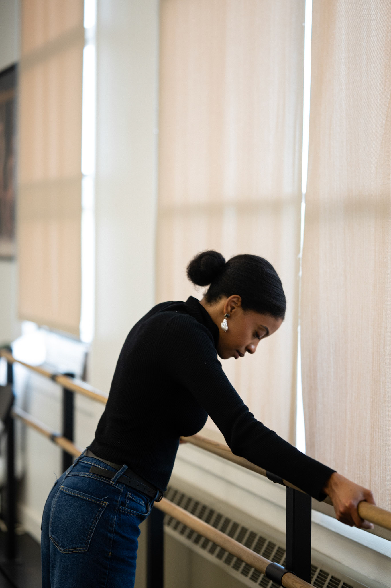 A woman in a black top and jeans leans on a ballet barre, gazing downward with soft lighting.