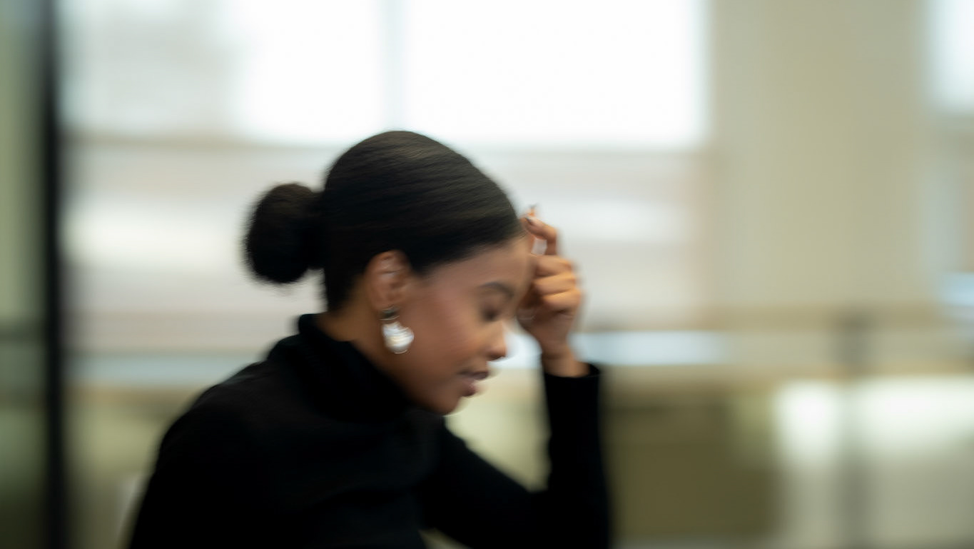 A woman in a black turtleneck with her hair in a bun, gently touching her forehead in a softly lit office setting