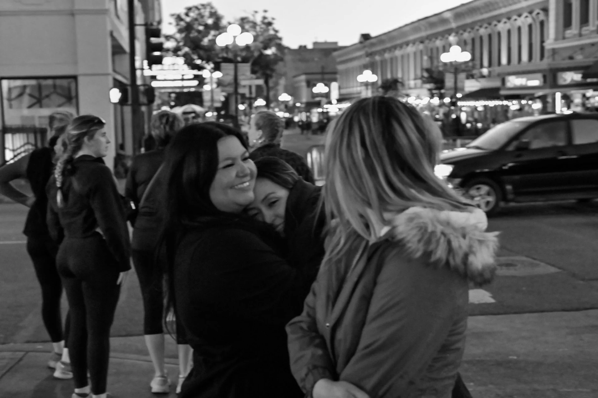 A black-and-white street scene capturing a warm, candid moment of friends embracing under city lights.