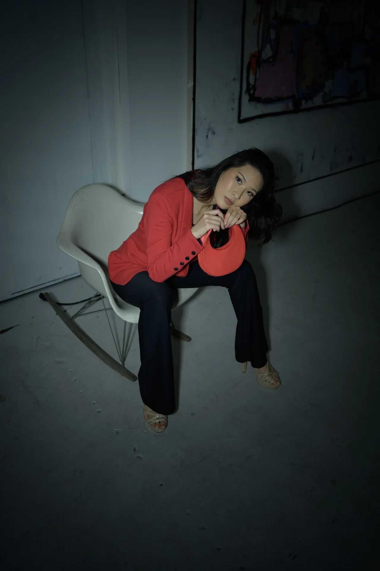 A woman in a red jacket sits on a white rocking chair in a dimly lit room, holding a red hat.