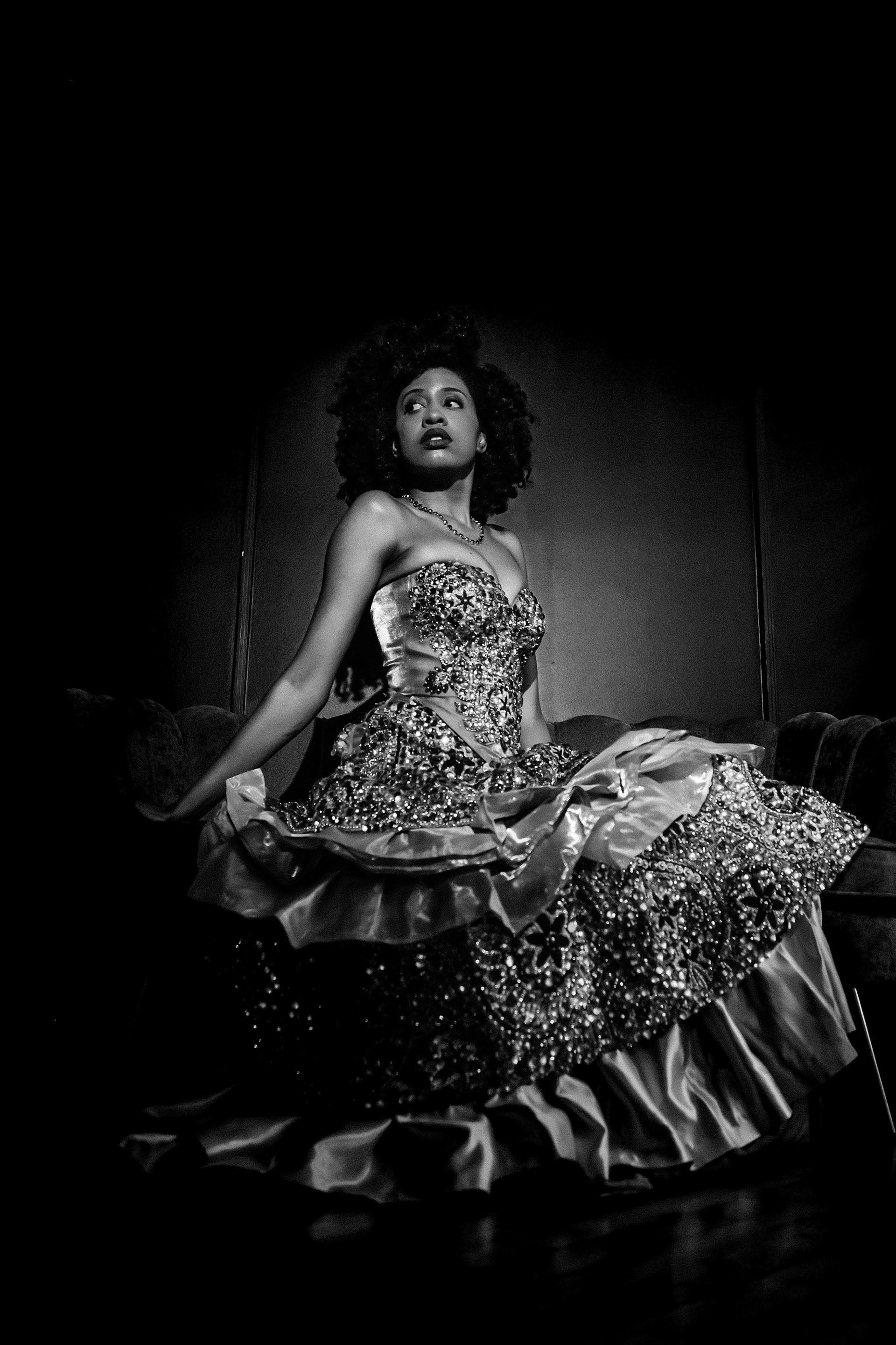 A woman in an ornate, beaded gown sits dramatically against a dark background, gazing upward.
