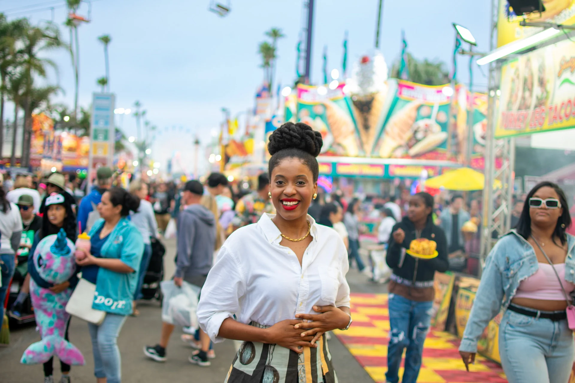 A vibrant carnival scene with a smiling woman in the foreground, surrounded by colorful stalls and bustling crowds.