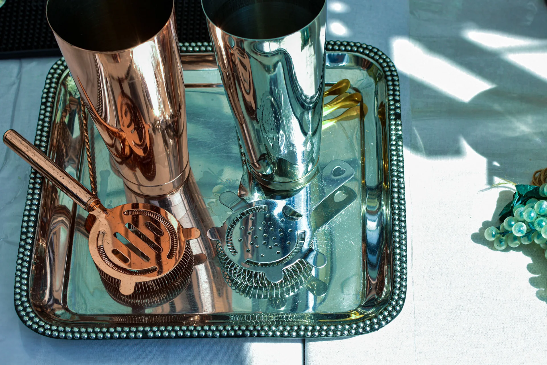 A gleaming set of bar tools rests elegantly on a beaded metal tray, bathed in bright sunlight.