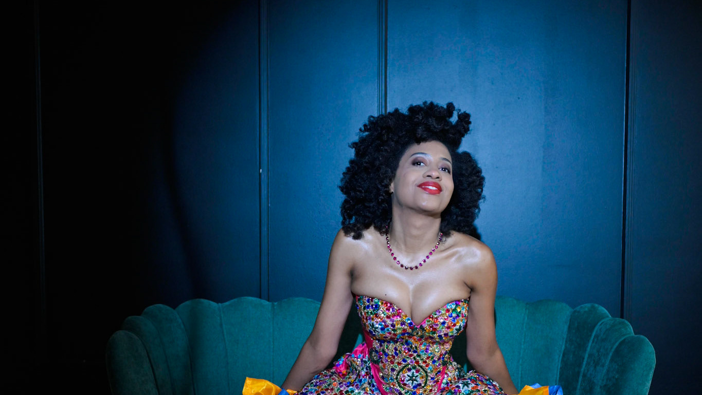A woman with voluminous curls sits on a teal couch, wearing a vibrant, beaded dress against a deep blue backdrop.