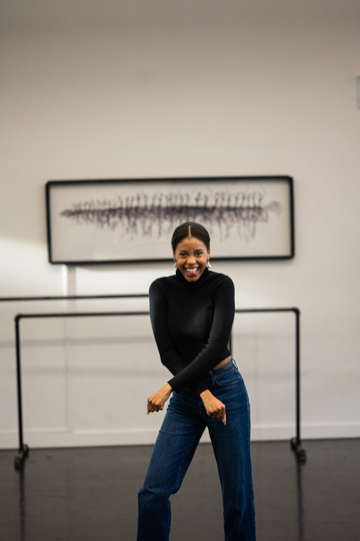 A woman smiles playfully in a dance studio with ballet bars and abstract art in the background.