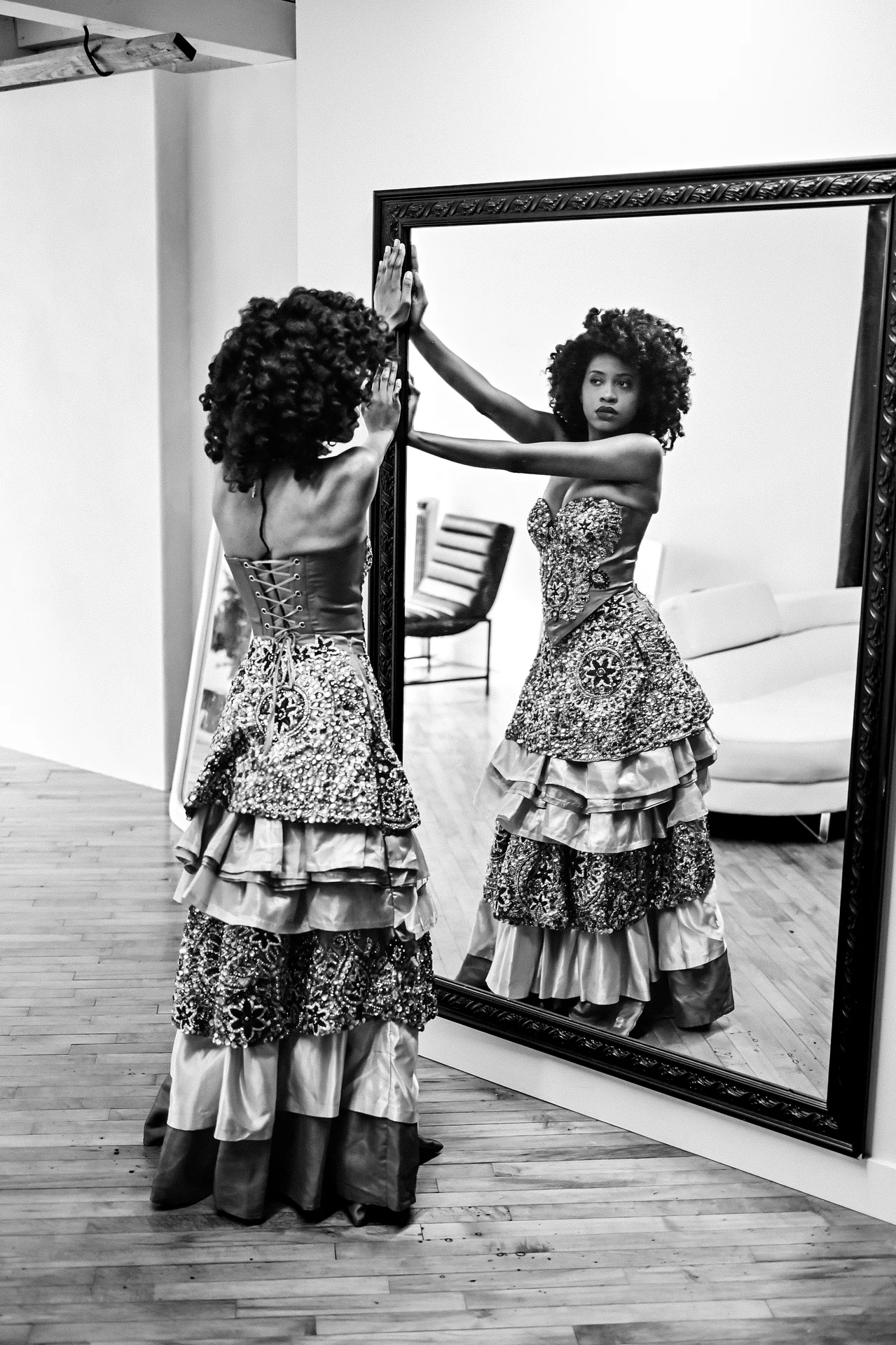 A woman in a blue velvet dress and black beret sits on a stool, gazing at herself in an arched mirror while adjusting her makeup.